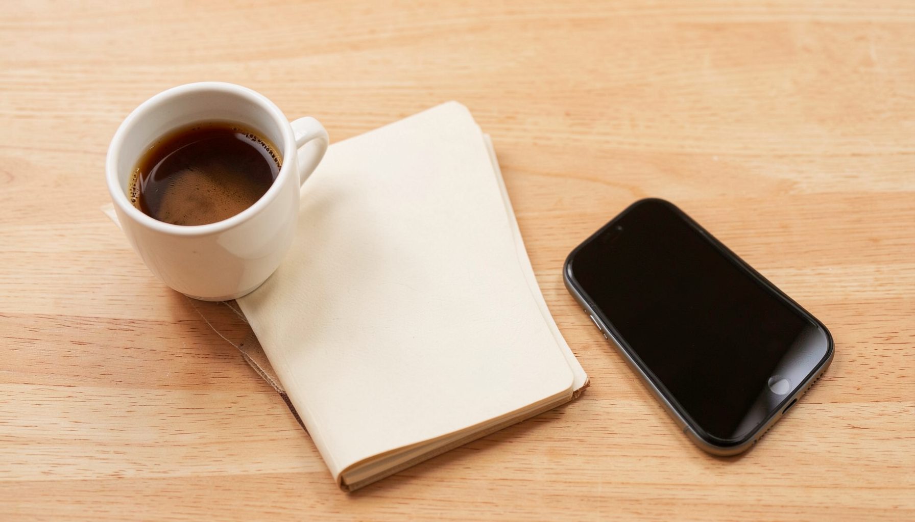 A warm-toned flat-lay of a notebook, a cup of coffee, and a smartphone on a light wood desk, evoking a calm and cozy mood